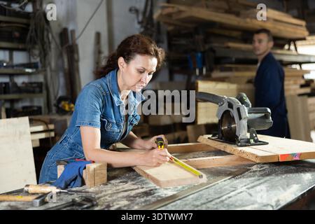 Work in woodworking enterprise - woman measures length of wooden board with tape measure Stock Photo