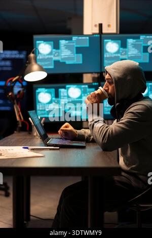Male programmer works late in office, sipping coffee while reviewing coding software on laptop. Caucasian man wearing hoodie, drinks caffeine and monitors encrypted data on digital device. Stock Photo
