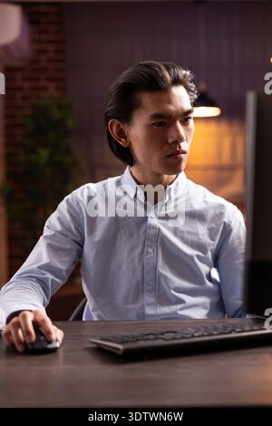Focused asian man works remotely at desk, using mouse to scroll through online documents, proofreading data. Professional male freelancer researches business strategies, improving results from home. Stock Photo