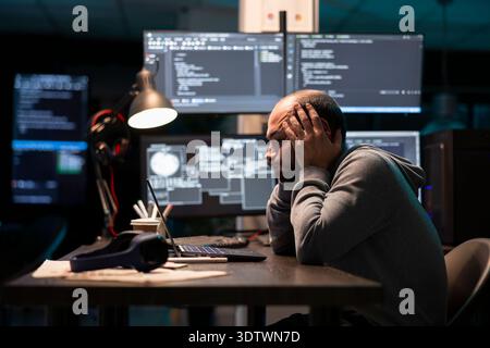 Frustrated software engineer leans forward, holding face in exhaustion and reviewing errors. Tired programmer works late at desk surrounded by coding interfaces and system windows. Stock Photo