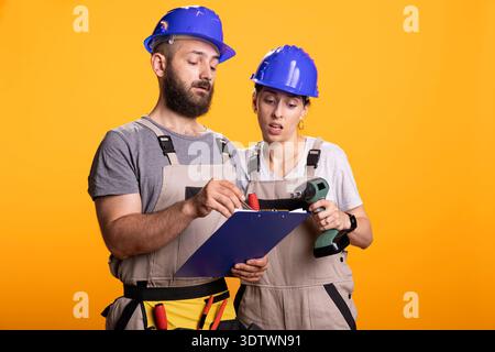 Male and female builders with clipboard, discussing upcoming tasks against isolated background. Caucasian coworkers in protective gear show teamwork and shared responsibility in construction planning. Stock Photo