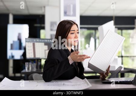 Focused female professional holds white architectural model while discussing with client on video call. Young asian architect evaluating project details with remote team at construction site. Stock Photo