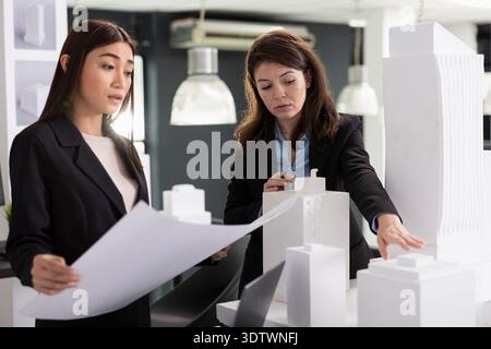 Two female architects evaluate white architectural model and review printed construction plans. Professional creative designers collaborating on building project decisions and execution timeline. Stock Photo