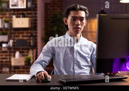 Focused businessman works on digital tasks, sitting at modern desk in home office. Handsome asian entrepreneur reviews marketing data and emails to improve small business strategies. Stock Photo