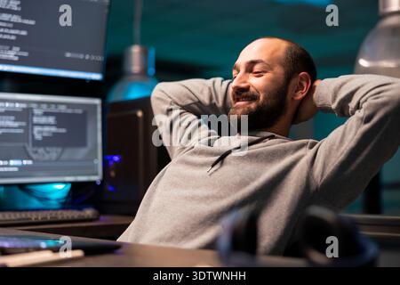 Relaxed programmer stretches back comfortably at workstation after finishing coding milestone. Smiling cybersecurity specialist feeling satisfied after handling demanding digital environment. Stock Photo