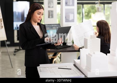 Professional designer reading digital blueprints and checking details on physical architectural model. Focused architect evaluating project requirements and creating reports for client presentations. Stock Photo