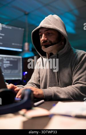 Hooded injured man working diligently on cybersecurity, analyzing networks and spyware risks. Focused male programmer with neck brace writing code and concentrating on improving company networks. Stock Photo