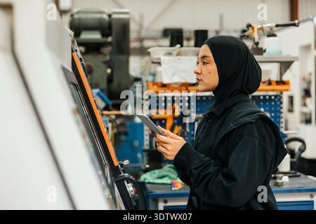 Young female worker wearing hijab operating machine with digital tablet in factory Stock Photo