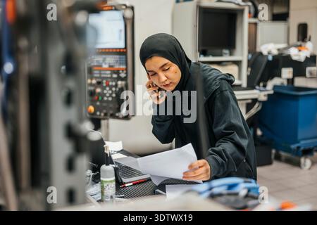 Young female employee talking on phone while reading document in metal factory Stock Photo