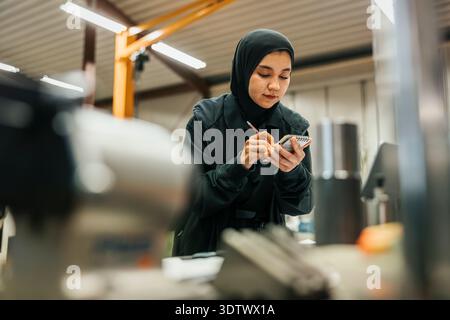 Low angle view of female factory worker wearing hijab taking notes on note pad Stock Photo