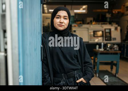 Portrait of confident female worker wearing hijab standing with hand in pocket near factory door Stock Photo