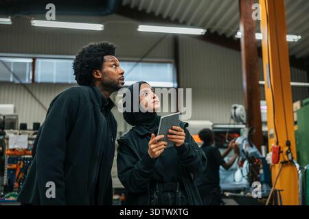 Male worker standing near female coworker operating machine with digital tablet in factory Stock Photo