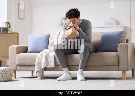 Young man with paper bag having panic attack on sofa at home Stock Photo