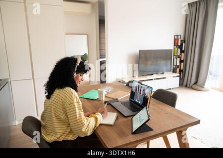 A woman with headphones participates in a video call at home, taking notes in a notebook. A laptop and tablet with charts enhance this remote work set Stock Photo