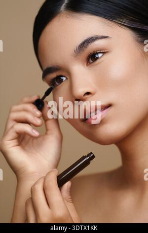 A young woman is applying mascara, enhancing her eyelashes for a stunning and captivating look. Her smooth skin and focused expression showcase beauty Stock Photo