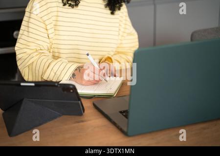 A woman in a striped t-shirt taking notes in a notebook while seated at a desk with a laptop and tablet, focusing on her hands, capturing a productive Stock Photo