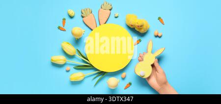 Hand with bunny-shaped cookie, blank card, Easter eggs, chicks and flowers on blue background, top view Stock Photo
