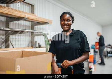 Portrait of smiling female worker standing near hand truck at warehouse Stock Photo