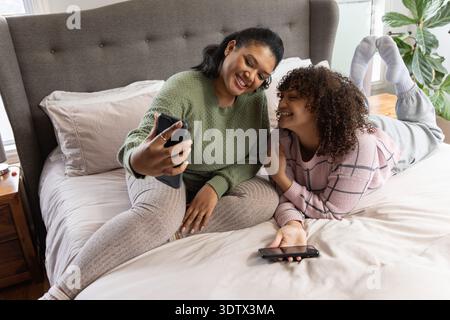 Female mother-daughter taking selfie on bed in knit sweaters holding phones smiling near window Stock Photo