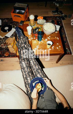 High angle low section view of young woman eating fast food with feet up on table at home Stock Photo