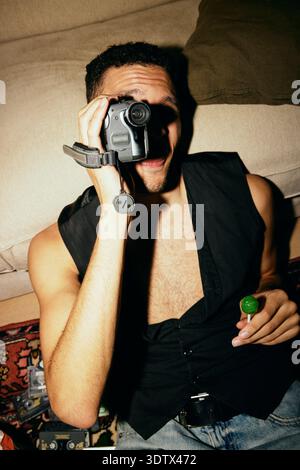 Young man holding candy while looking through vintage video camera at home Stock Photo