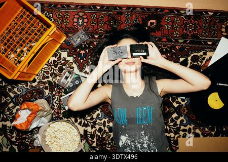 Directly above shot of woman covering eyes with audio cassettes while lying on carpet at home Stock Photo
