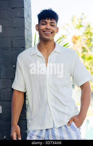 African American man mid-20s leaning on dark-gray column by pool in white shirt blue striped shorts Stock Photo