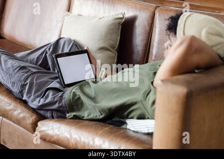 Tablet with blank white screen is resting on brown leather sofa at home, with folded blanket Stock Photo