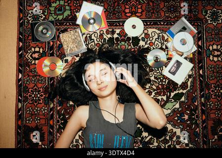 Smiling young woman listening to music through headphones while lying on carpet amidst CDs Stock Photo