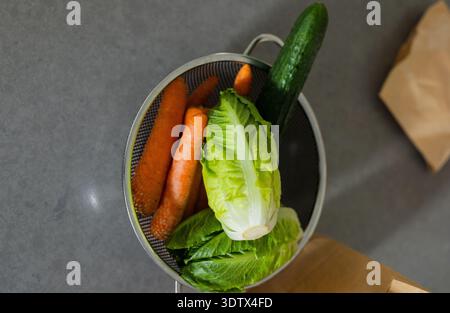 Metal colander holding three carrots, romaine and cucumber on dark gray countertop near paper bag Stock Photo