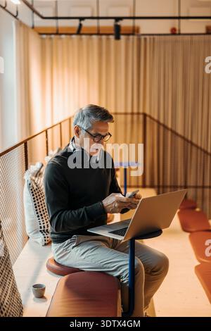 Senior businessman using smart phone while sitting on couch with laptop at modern coworking space Stock Photo