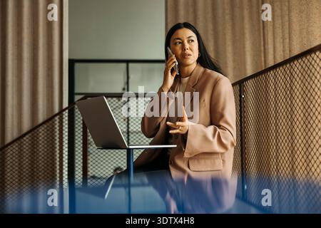 Busy female entrepreneur talking on smart phone while sitting with laptop at office Stock Photo