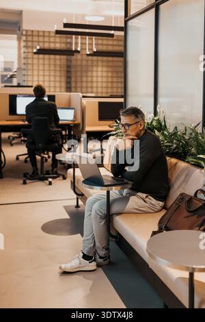Full length of thoughtful senior businessman looking at laptop while sitting on seat at office Stock Photo