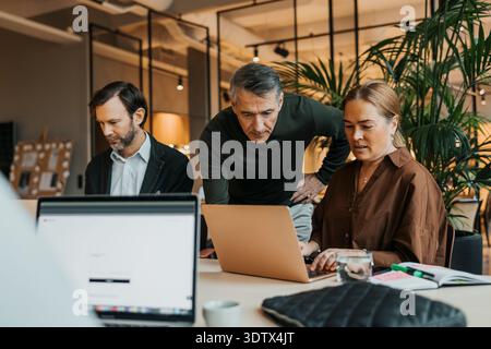 Senior male entrepreneur assisting female coworker using laptop while working at desk in office Stock Photo