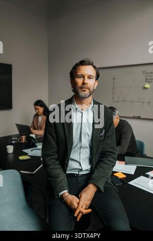 Portrait of confident businessman sitting on table in office meeting room Stock Photo