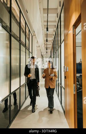 Full length of male and female entrepreneurs talking while walking in illuminated office corridor Stock Photo