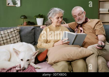 Senior Caucasian Couple Sitting on Sofa Reading Book Together at Home Stock Photo