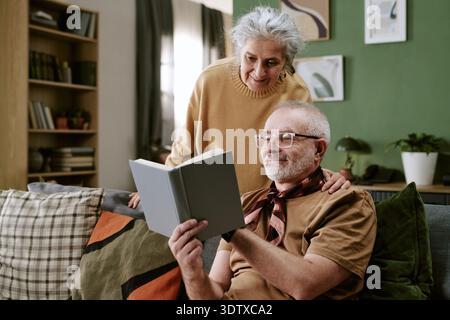 Senior Caucasian Man Reading Book While Senior Caucasian Woman Standing Behind Stock Photo
