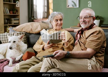Senior Caucasian Couple Relaxing on Sofa with Dog and Reading Book Together Stock Photo