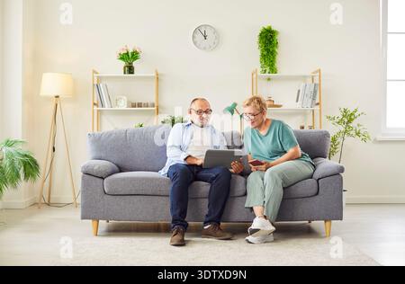 Senior couple sitting on couch at home using tablet and smartphone together Stock Photo