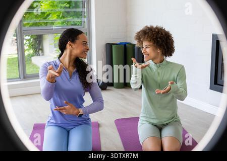 African American female friends in gymwear sitting on purple mats, smiling, gesturing by window Stock Photo