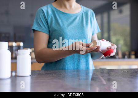 White pill bottle tipping tablet into palm on stone countertop in kitchen with two bottles Stock Photo