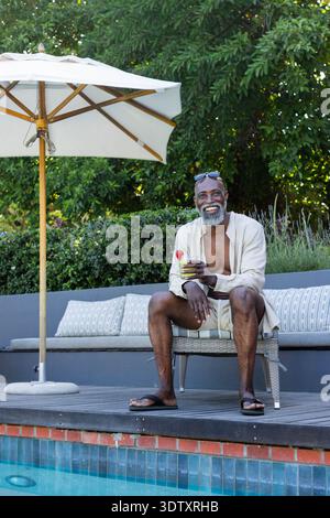 Senior African man sitting on wicker bench at pool deck wearing light shirt, holding cocktail glass Stock Photo