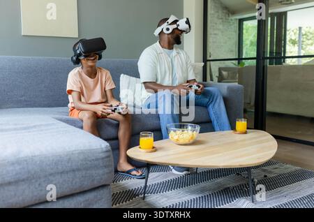African American father and teenage son wearing VR headsets on gray sofa, holding controllers Stock Photo