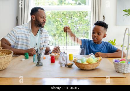 African American father guiding child son while painting eggs at dining table with paint jars Stock Photo