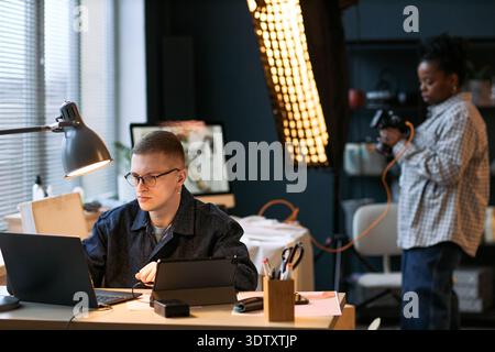 Caucasian young adult man working on laptop at desk, Black young adult woman standing in background holding professional camera in modern office studio setting Stock Photo