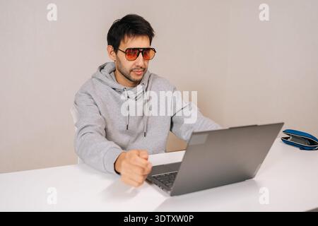 Portrait of young man wearing stylish blue light blocking glasses and hoodie, sitting at desk and typing on laptop, concentrating on screen while Stock Photo