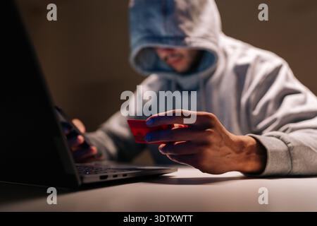 Portrait of hooded unrecognizable person in dark room holding phone and credit card while using laptop, depicting online fraud, identity theft and Stock Photo