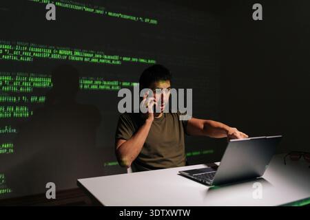 Busy programmer male sitting at desk with laptop, deeply engaged in phone conversation while analyzing projected green code, symbolizing complex Stock Photo