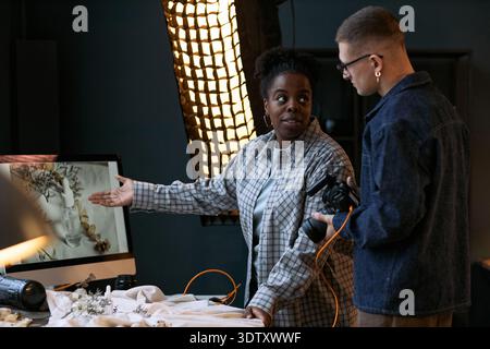 Black young adult woman explaining creative project to Caucasian young adult man holding camera, both standing near computer monitor displaying floral arrangement in studio setting Stock Photo